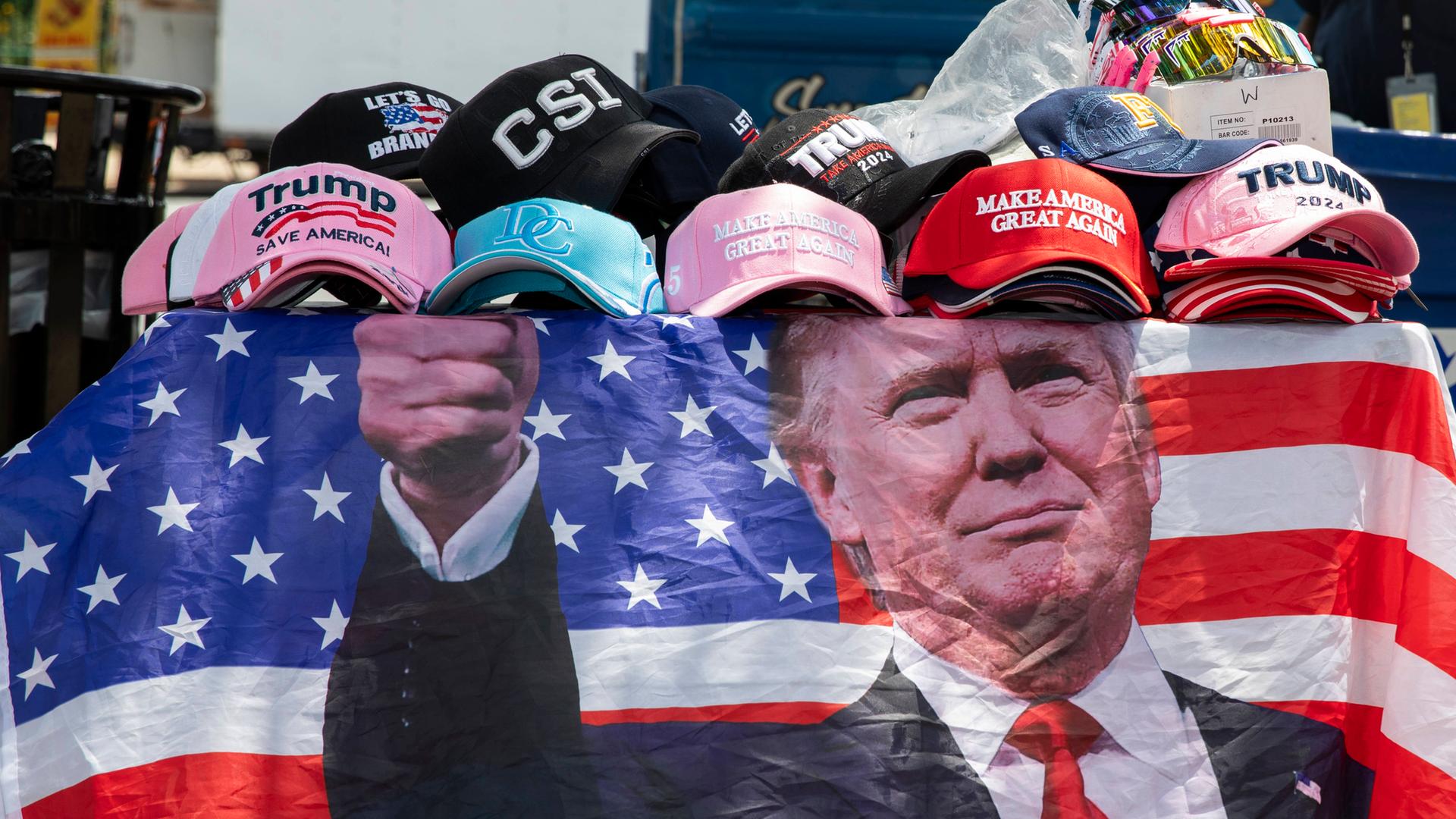 Eine mit dem Konterfei von Donald Trump versehene Fahne hängt an einem Verkaufsstand in Washington DC. Darauf liegen Basball-Mützen. Unter anderem auch die rote Mütze mit der Trump-Aufschrift: "Make America great again". Juni 29, 2024.