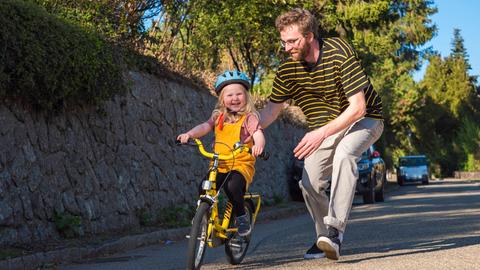 Ein Vater läuft auf einer Straße seiner Tochter mit ihrem Fahrrad hinterher. (Symbolbild)