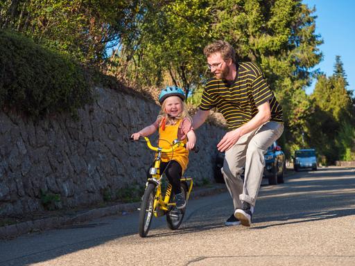 Ein Vater läuft auf einer Straße seiner Tochter mit ihrem Fahrrad hinterher. (Symbolbild)