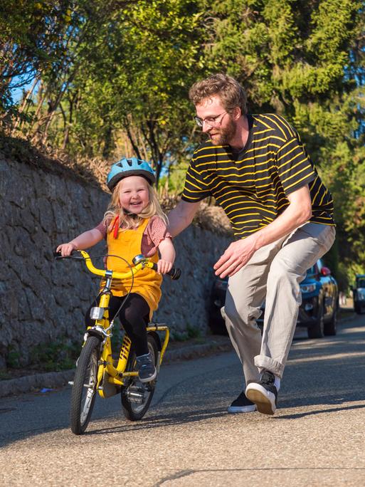 Ein Vater läuft auf einer Straße seiner Tochter mit ihrem Fahrrad hinterher. (Symbolbild)