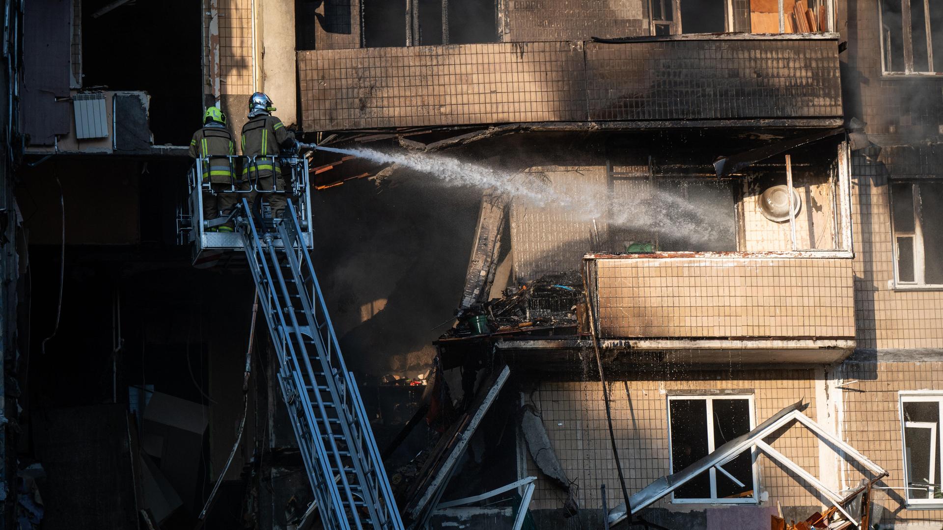 Zwei Feuerwehrleute stehen auf einer Drehleiter und löschen ein Feuer in einem Wohnhaus, das durch den Angriff russischer Drohnen verursacht wurde.