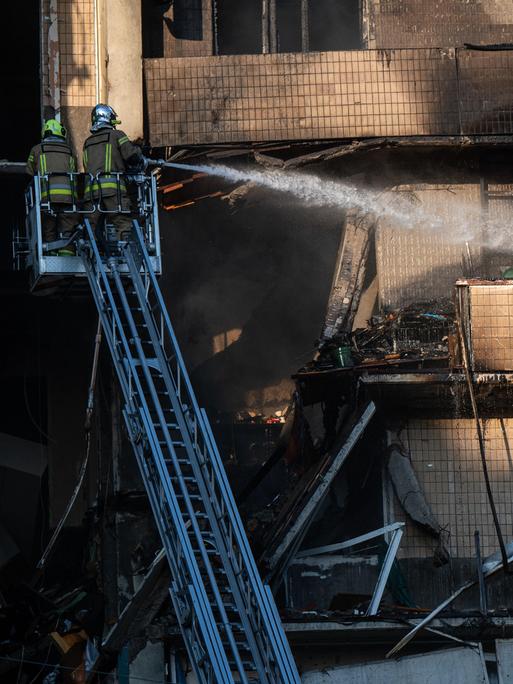 Zwei Feuerwehrleute stehen auf einer Drehleiter und löschen ein Feuer in einem Wohnhaus, das durch den Angriff russischer Drohnen verursacht wurde.