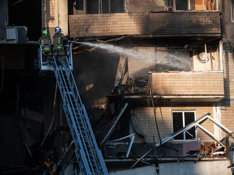 Zwei Feuerwehrleute stehen auf einer Drehleiter und löschen ein Feuer in einem Wohnhaus, das durch den Angriff russischer Drohnen verursacht wurde.