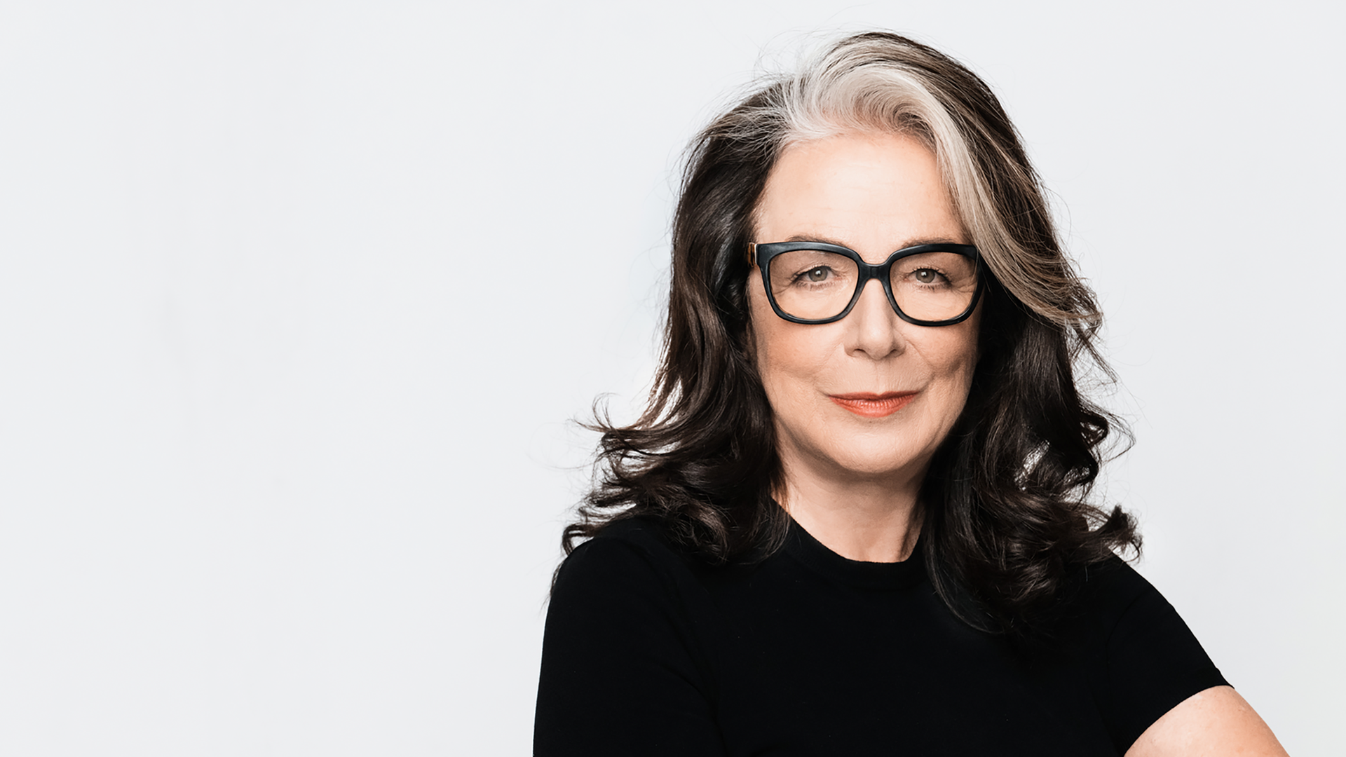 Evelyn Roll trägt eine schwarz umrandete Brille und ein schwarzes Oberteil. Sie sitzt vor einem weißen Hintergrund und schaut freundlich in die Kamera.