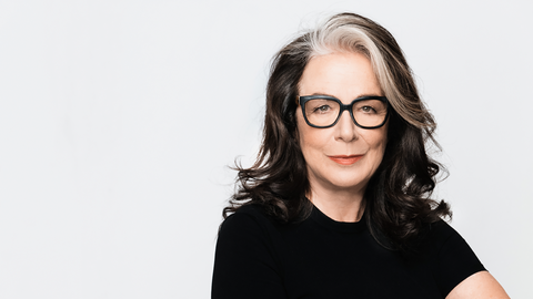 Evelyn Roll trägt eine schwarz umrandete Brille und ein schwarzes Oberteil. Sie sitzt vor einem weißen Hintergrund und schaut freundlich in die Kamera.