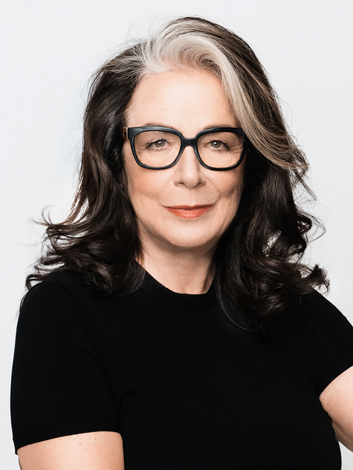 Evelyn Roll trägt eine schwarz umrandete Brille und ein schwarzes Oberteil. Sie sitzt vor einem weißen Hintergrund und schaut freundlich in die Kamera.