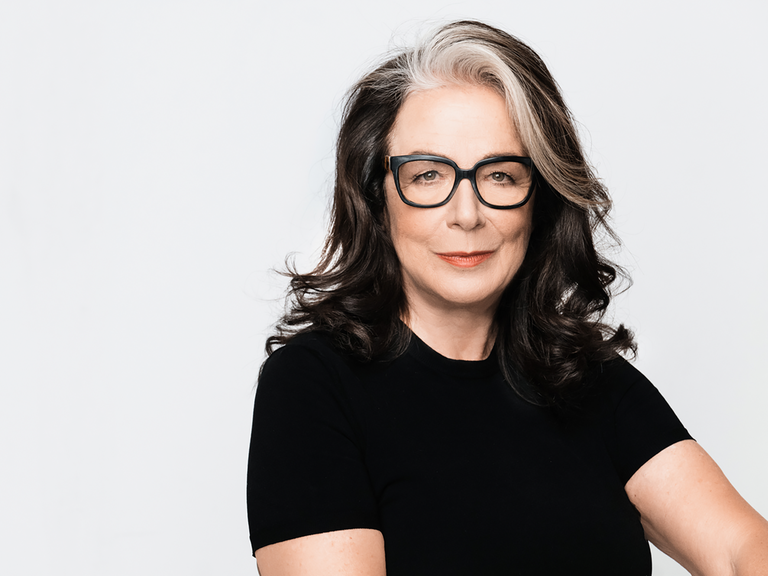 Evelyn Roll trägt eine schwarz umrandete Brille und ein schwarzes Oberteil. Sie sitzt vor einem weißen Hintergrund und schaut freundlich in die Kamera.