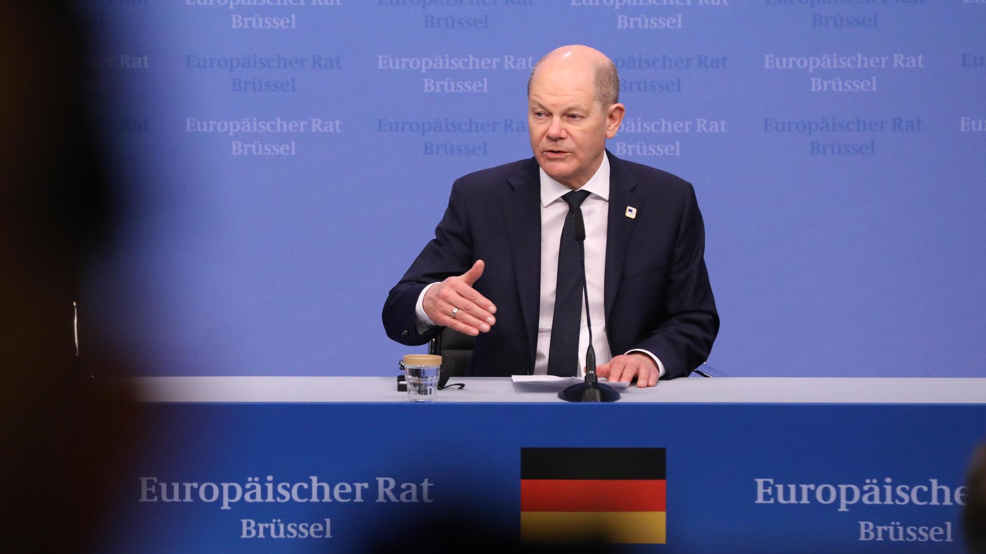 Ein glatzköpfiger Mann, Bundeskanzler Olaf Scholz, steht vor blauem Hintergrund an einem Rednerpult mit Mikrofon.