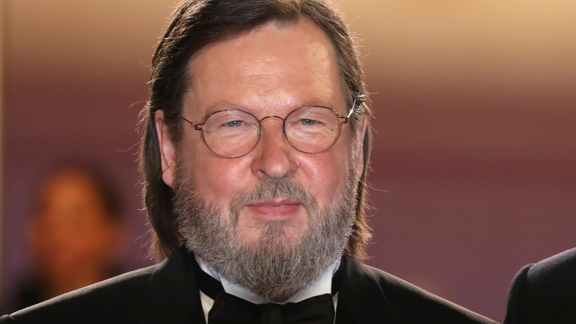 Der Regisseur Lars von Trier mit Brille, Bart und längeren Haaren trägt eine Fliege, weißes Hemd und Jackett.