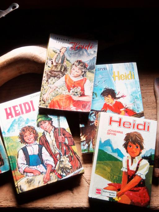 Fünf verschiedene Ausgaben des Kinderbuchs „Heidi“ liegen auf einem Tiesch. Daneben liegen geschnitzte Holzlöffel. Die Cover-Motive der Bücher wirken volkstümlich mit romantisierter Alpenkulisse. 