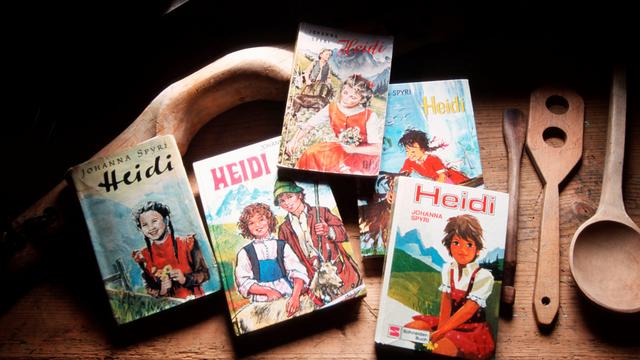 Fünf verschiedene Ausgaben des Kinderbuchs „Heidi“ liegen auf einem Tiesch. Daneben liegen geschnitzte Holzlöffel. Die Cover-Motive der Bücher wirken volkstümlich mit romantisierter Alpenkulisse. 