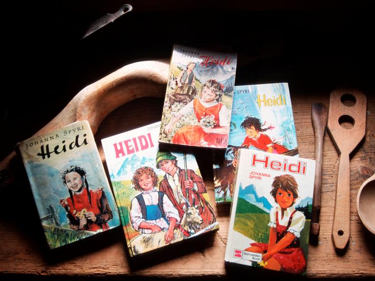 Fünf verschiedene Ausgaben des Kinderbuchs „Heidi“ liegen auf einem Tiesch. Daneben liegen geschnitzte Holzlöffel. Die Cover-Motive der Bücher wirken volkstümlich mit romantisierter Alpenkulisse. 