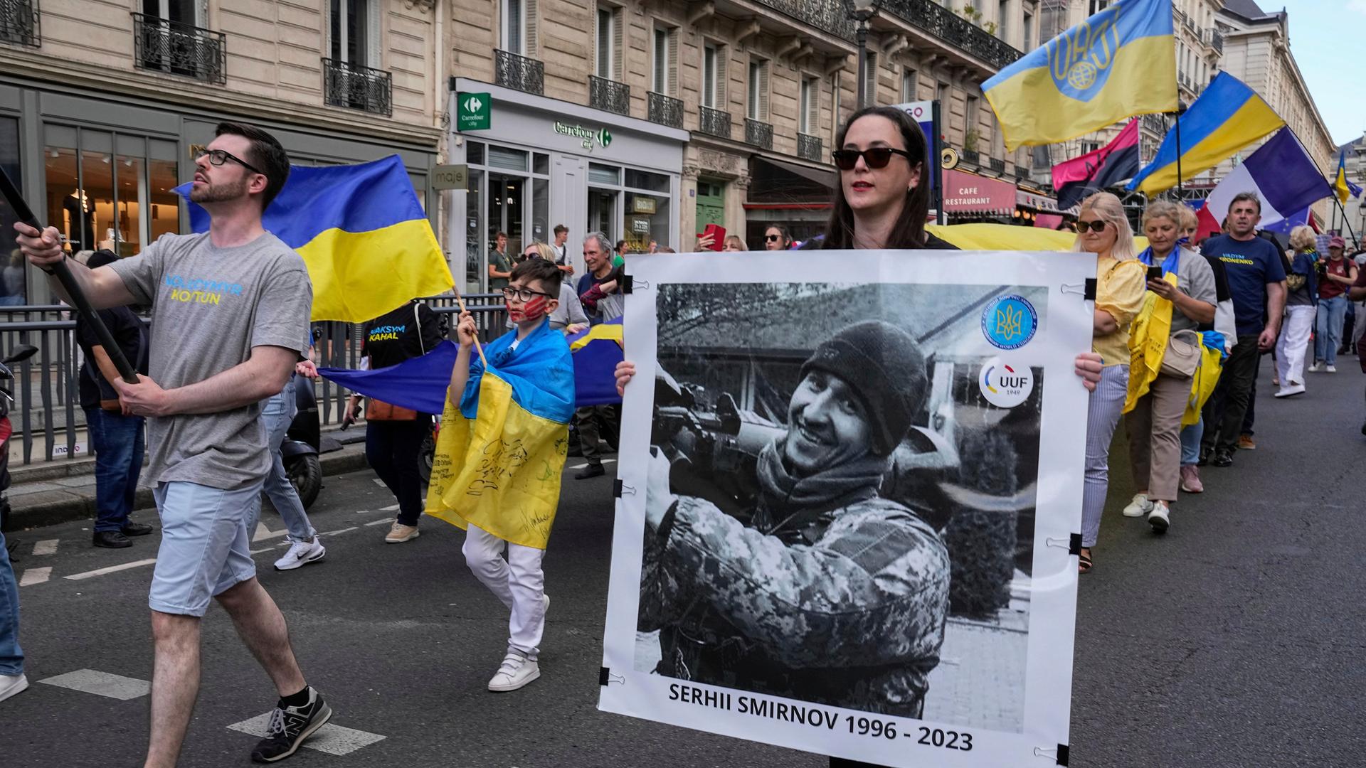 Menschen ziehen mit ukrainischen Flaggen durch die französische Hauptstadt Paris. Eine Frau hält ein großes Foto mit dem Bild eines ukrainischen Sportlers. Er trägt eine Uniform. Unter dem Foto stehen die Jahreszahlen 1996 bis 2023. 