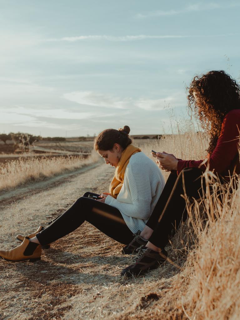 Zwei Frauen sitzen mit Mobiltelefonen in der Hand an einem Feldweg. 