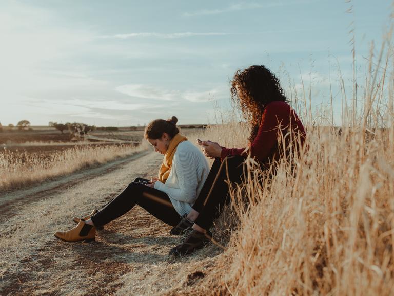 Zwei Frauen sitzen mit Mobiltelefonen in der Hand an einem Feldweg. 