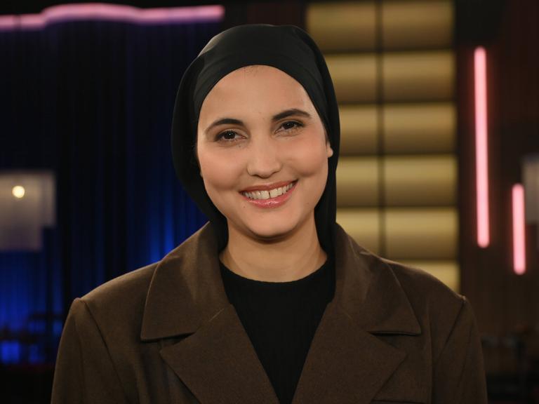 Comedienne Anissa Loucif zu Gast in der WDR Talkshow Kölner Treff. Sie trägt ein dunkles Kopftuch und lacht vergnügt in die Kamera. 
