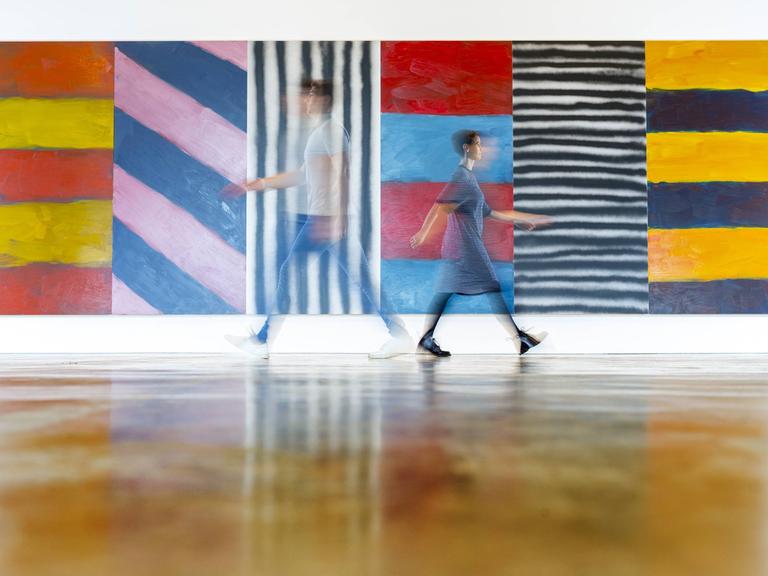 Kerry Chase und Sam Whitworth laufen an einem Gemälde von Sean Scully entlang mit dem Titel "Blue Note". Man sieht beide nur schemenhaft.