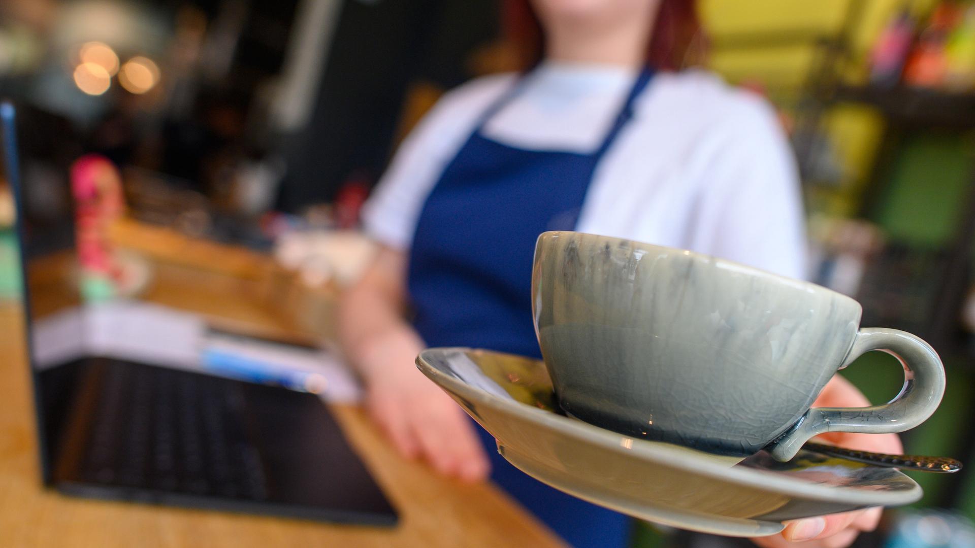 Eine Bedienung bringt in einem Cafe eine Tasse Kaffee an den Tisch. Die Bedienung ist nur verschwommen erkennbar, die Kaffeetasse im Vordergrund steht im Fokus. Eine Bedienung bringt in einem Cafe eine Tasse Kaffee an den Tisch. Die Bedienung ist nur verschwommen erkennbar, die Kaffeetasse im Vordergrund steht im Fokus.