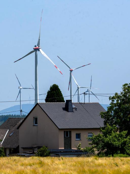 Mehrere Windkraftanlagen erheben sich hinter den Dächern von Wohnhäusern in einer ländlichen Landschaft. Im Vordergrund ein reifes Getreidefeld, dahinter Bäume und Hecken, im Hintergrund bewaldete Hügel unter blauem Himme Mehrere Windkraftanlagen erheben sich hinter den Dächern von Wohnhäusern in einer ländlichen Landschaft. Im Vordergrund ein reifes Getreidefeld, dahinter Bäume und Hecken, im Hintergrund bewaldete Hügel unter blauem Himme