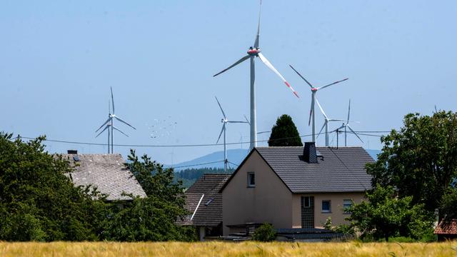 Mehrere Windkraftanlagen erheben sich hinter den Dächern von Wohnhäusern in einer ländlichen Landschaft. Im Vordergrund ein reifes Getreidefeld, dahinter Bäume und Hecken, im Hintergrund bewaldete Hügel unter blauem Himme