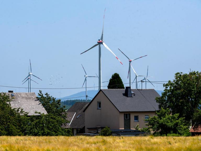 Mehrere Windkraftanlagen erheben sich hinter den Dächern von Wohnhäusern in einer ländlichen Landschaft. Im Vordergrund ein reifes Getreidefeld, dahinter Bäume und Hecken, im Hintergrund bewaldete Hügel unter blauem Himme Mehrere Windkraftanlagen erheben sich hinter den Dächern von Wohnhäusern in einer ländlichen Landschaft. Im Vordergrund ein reifes Getreidefeld, dahinter Bäume und Hecken, im Hintergrund bewaldete Hügel unter blauem Himme
