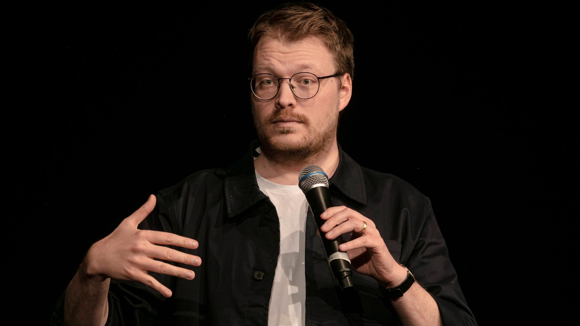 Maxi Gstettenbauer, ein Mann mittleren Alters mit runder Brille, dunkelblondem Haar und Dreitagebart steht auf einer Bühne.