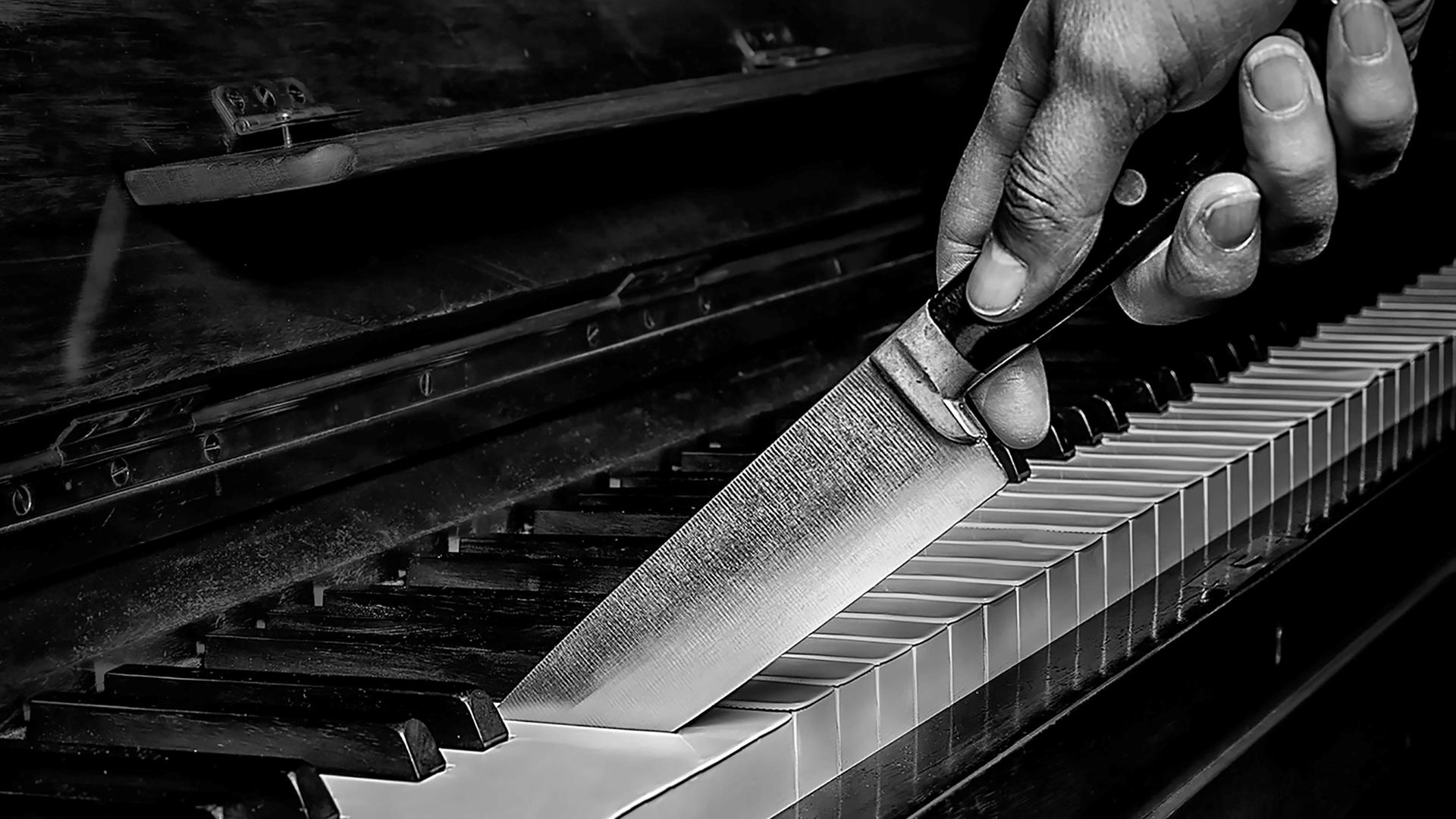 Schwarz-Weiß-Bild einer Hand mit einem großen Messer, das zwischen die Tasten eines Klaviers schneidet. Schwarz-Weiß-Bild einer Hand mit einem großen Messer, das zwischen die Tasten eines Klaviers schneidet.