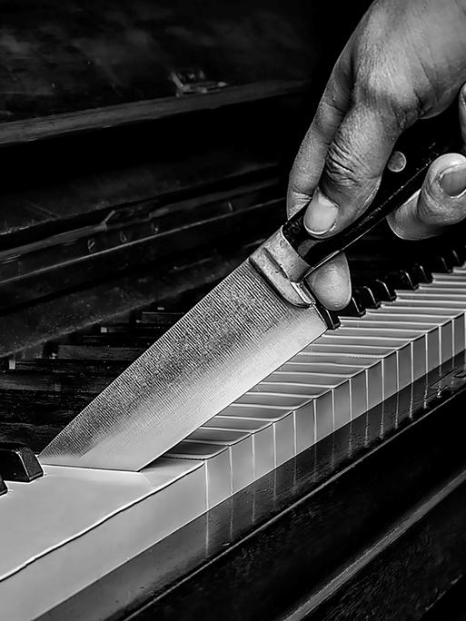 Schwarz-Weiß-Bild einer Hand mit einem großen Messer, das zwischen die Tasten eines Klaviers schneidet.