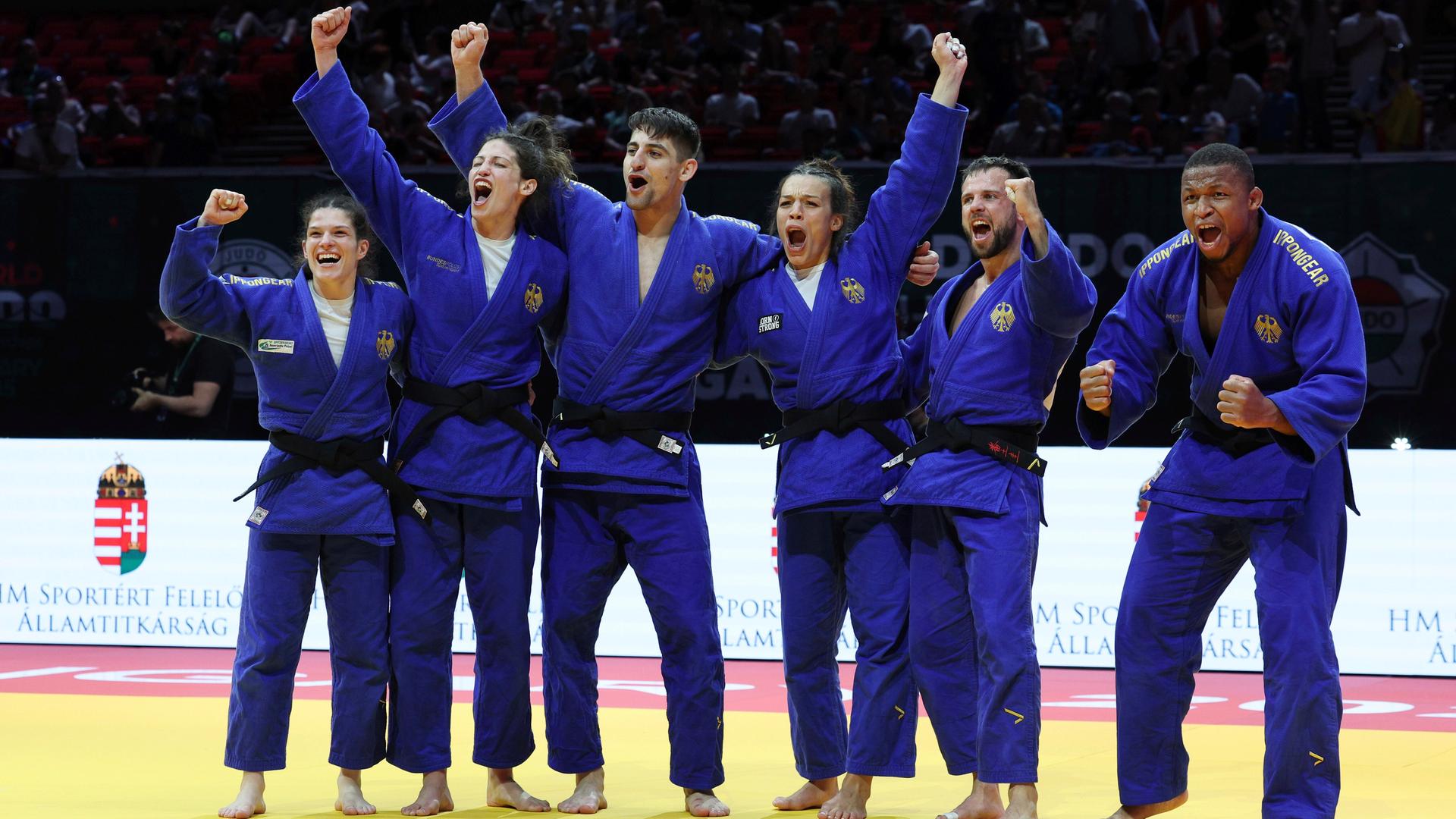 Das deutsche Team freut sich über die Bronze-Medaille. Die Sportlerinnen und Sportler stehen in blauen Judo-Anzügen nebeneinander und strecken die Hände in die Luft.