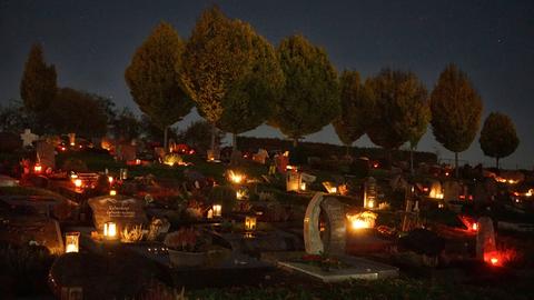 Wir sehen einen Friedhof bei Nacht, der von Grablichtern erleuchtet ist. 