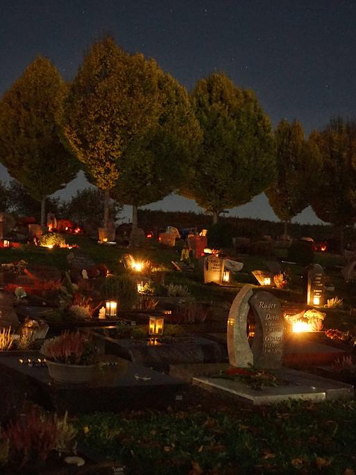 Wir sehen einen Friedhof bei Nacht, der von Grablichtern erleuchtet ist. 
