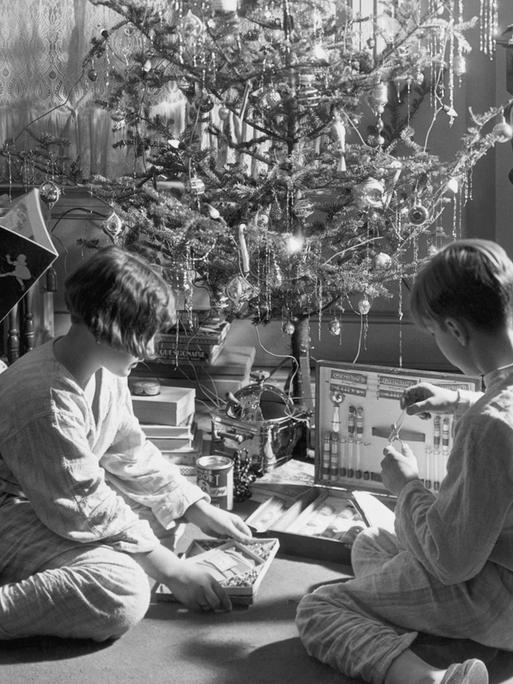 Historisches Schwarzweißfoto dreier Kinder, die um 1910 unter einem geschmückten Weihnachtsbaum sitzen und mit ihren Geschenken spielen.