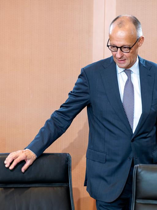 Bundeskanzler Friedrich Merz trägt einen Anzug und Krawatte und hat seine Hand an einem Stuhl. Er befindet sich dabei in einem Raum im Bundeskanzleramt.