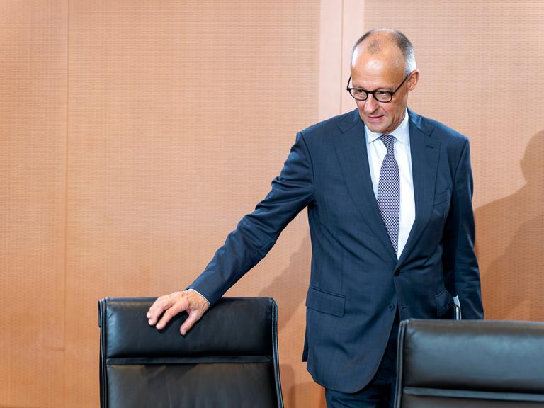 Bundeskanzler Friedrich Merz trägt einen Anzug und Krawatte und hat seine Hand an einem Stuhl. Er befindet sich dabei in einem Raum im Bundeskanzleramt.