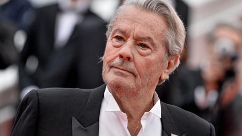 Alain Delon im schwarzen Anzug, mit offenem Hemdkragen und grauen Haaren. Er schaut auf dem roten Teppich beim Cannes Filmfestival zur Seite.