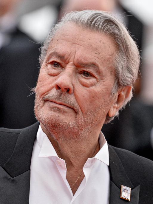 Alain Delon im schwarzen Anzug, mit offenem Hemdkragen und grauen Haaren. Er schaut auf dem roten Teppich beim Cannes Filmfestival zur Seite.