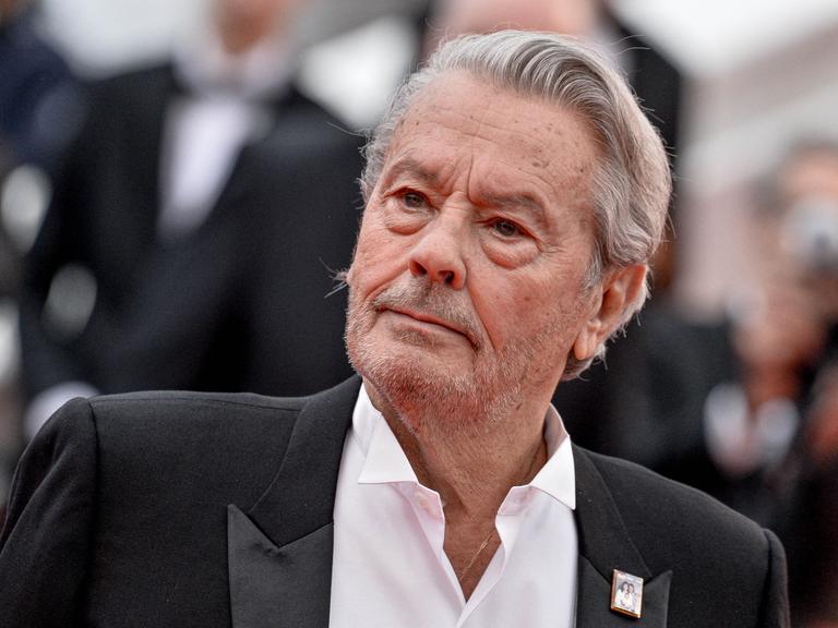 Alain Delon im schwarzen Anzug, mit offenem Hemdkragen und grauen Haaren. Er schaut auf dem roten Teppich beim Cannes Filmfestival zur Seite.