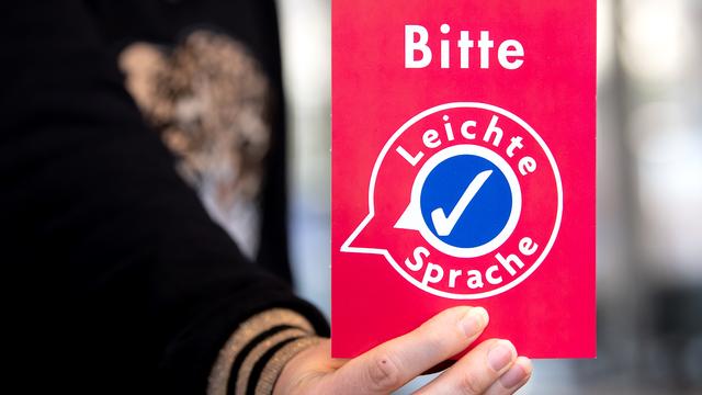 Eine Hand hält eine Rote Karte mit weißer Schrift hoch. Darauf steht: Bitte Leichte Sprache.