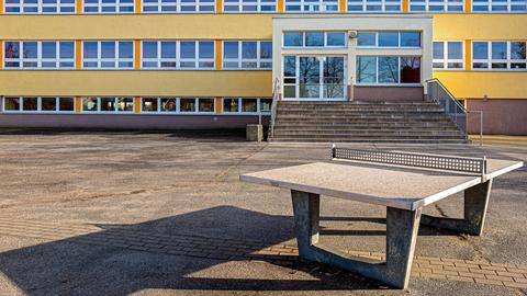 Das Bild zeigt einen leeren Schulhof mit Schulgebäude und einer Tischtennisplatte im Vordergrund