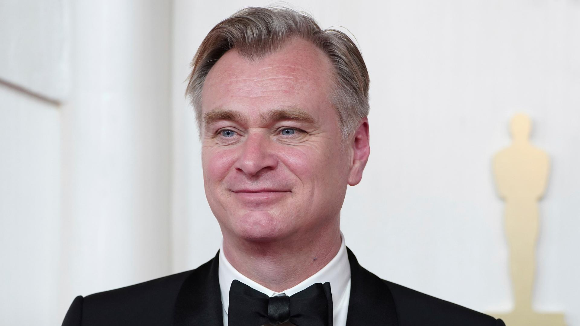 Christopher Nolan vor einer weißen Wand. Er lächelt. Im Hintergrund ist die Silhouette einer Oscar-Statue zu sehen. Christopher Nolan vor einer weißen Wand. Er lächelt. Im Hintergrund ist die Silhouette einer Oscar-Statue zu sehen.
