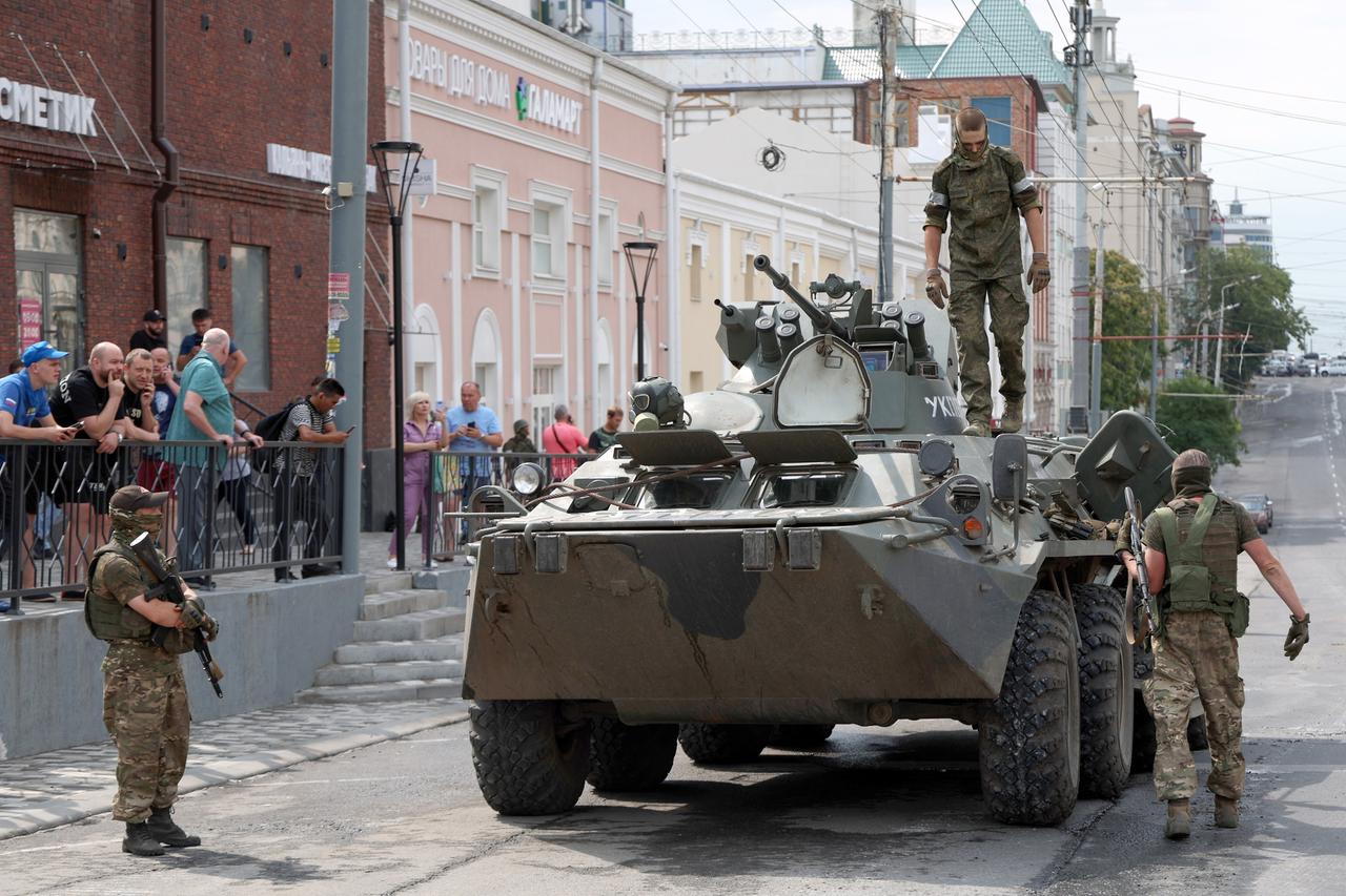 Drei Männer in Militärkleidung sind um einen Panzer zu sehen, am Rand stehen Zivilisten.