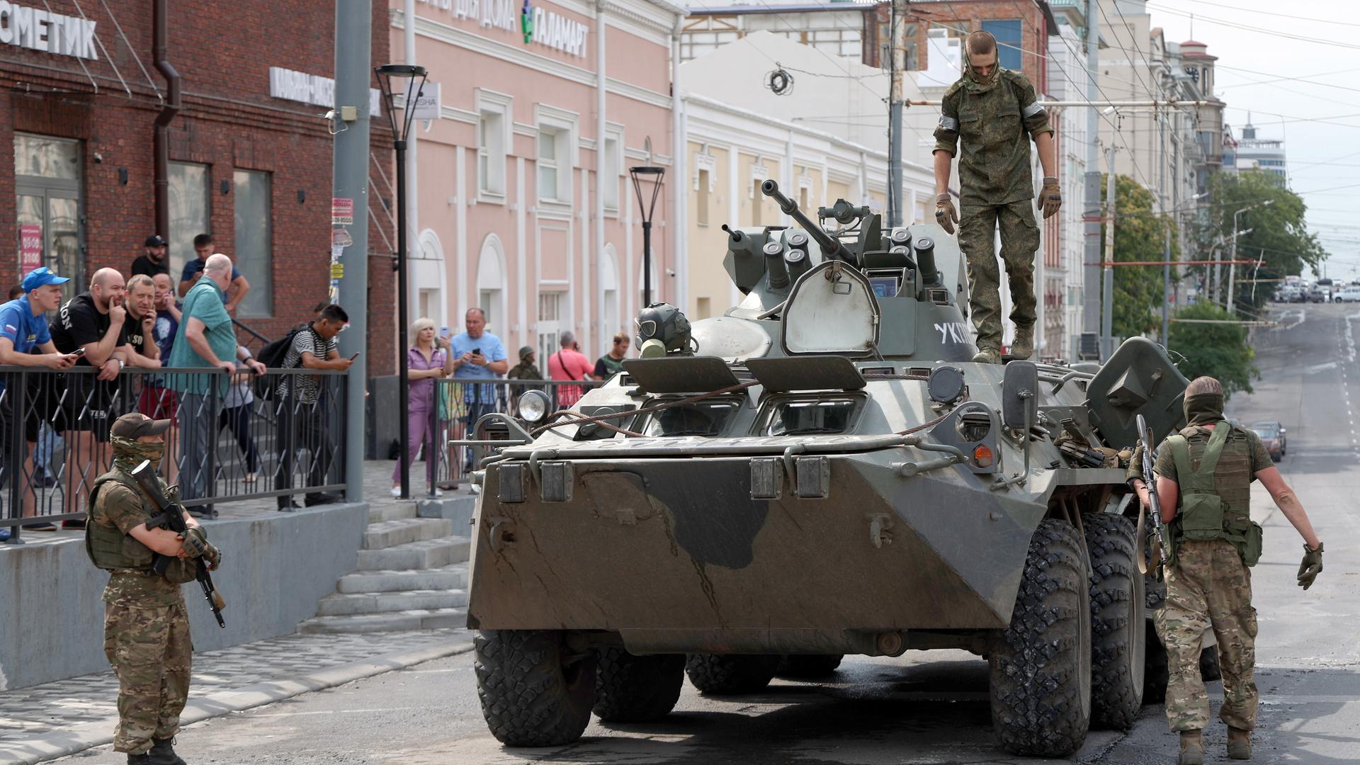 Drei Männer in Militärkleidung sind um einen Panzer zu sehen, am Rand stehen Zivilisten.