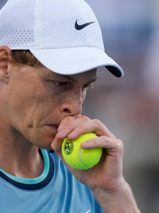 Tennis-Spieler Jannik Sinner in einer Proträt-Aufnahme hält sich einen Tennisball während eines Turnierspiels nachdenkend ans Gesicht.