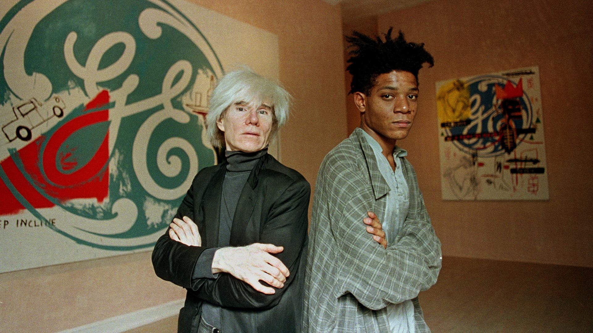 Die Künstler Andy Warhol (links) und Jean-Michel Basquiat stehen Rücken an Rücken vor ihren gemeinsamen Kunstwerken.