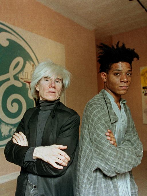 Die Künstler Andy Warhol (links) und Jean-Michel Basquiat stehen Rücken an Rücken vor ihren gemeinsamen Kunstwerken.