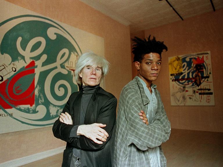 Die Künstler Andy Warhol (links) und Jean-Michel Basquiat stehen Rücken an Rücken vor ihren gemeinsamen Kunstwerken.