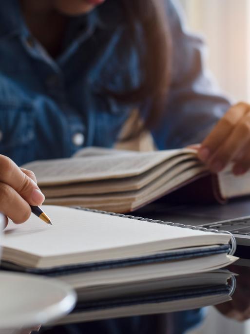Eine Frau liest ein Buch und macht sich daneben Notizen in einem Heft mit leeren Seiten.