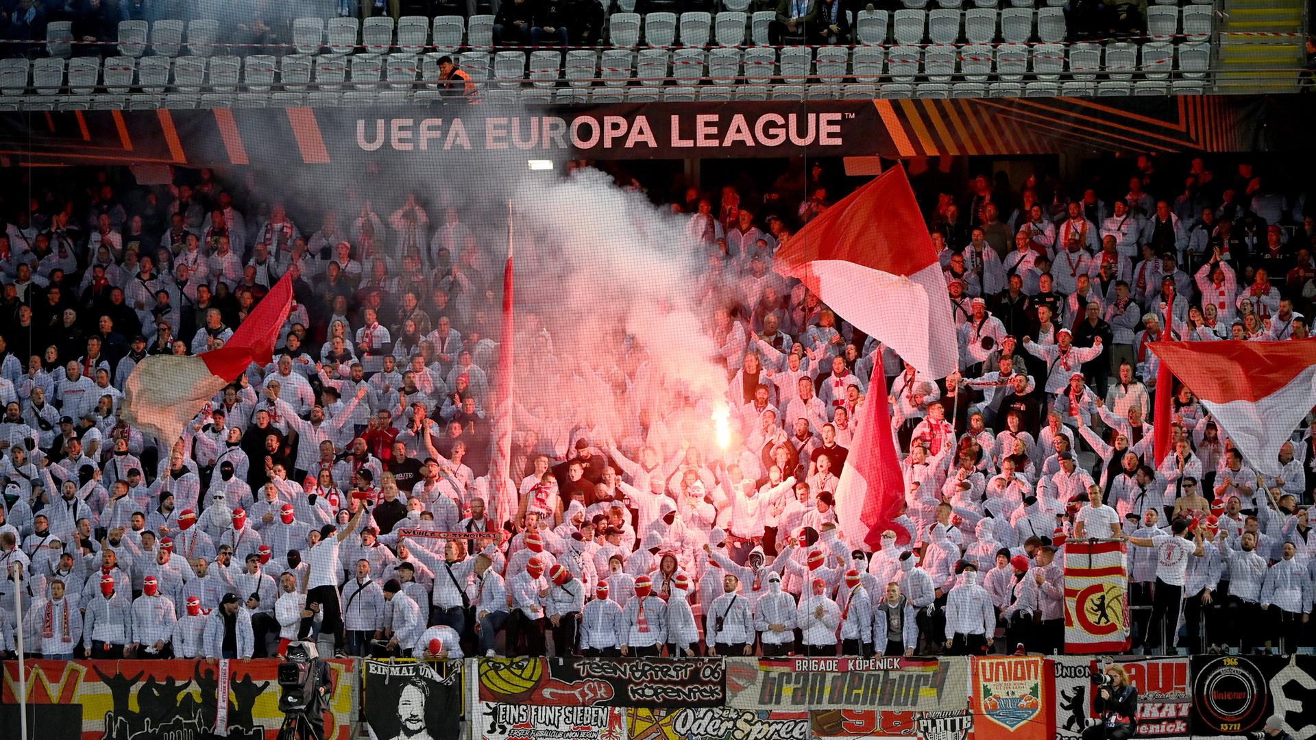 Fans stehen im Stadion und brennen Feuerwerk ab, es ist rotes Licht und Rauch zu sehen.