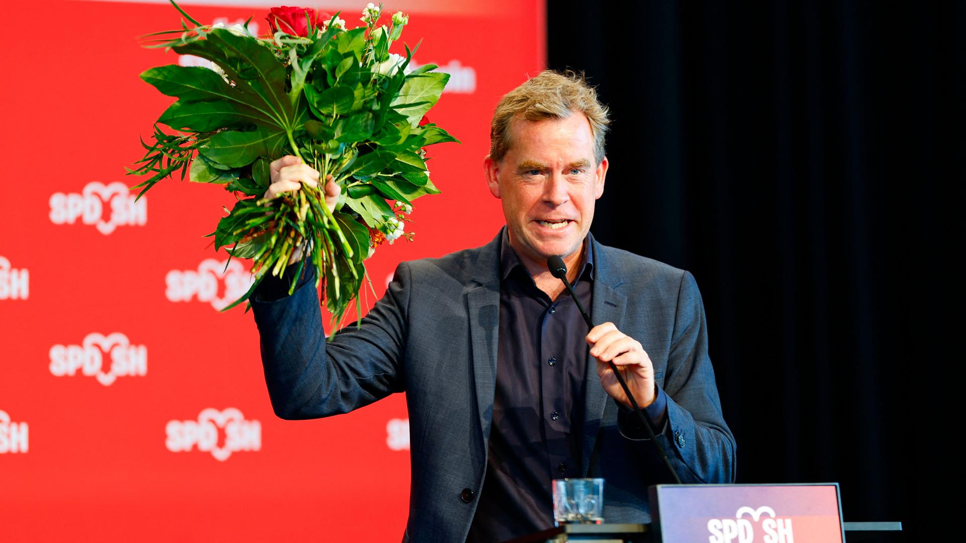 Ulf Kämpfer jubelt mit einem Blumenstrauß in der Hand auf einer SPD-Veranstaltung.