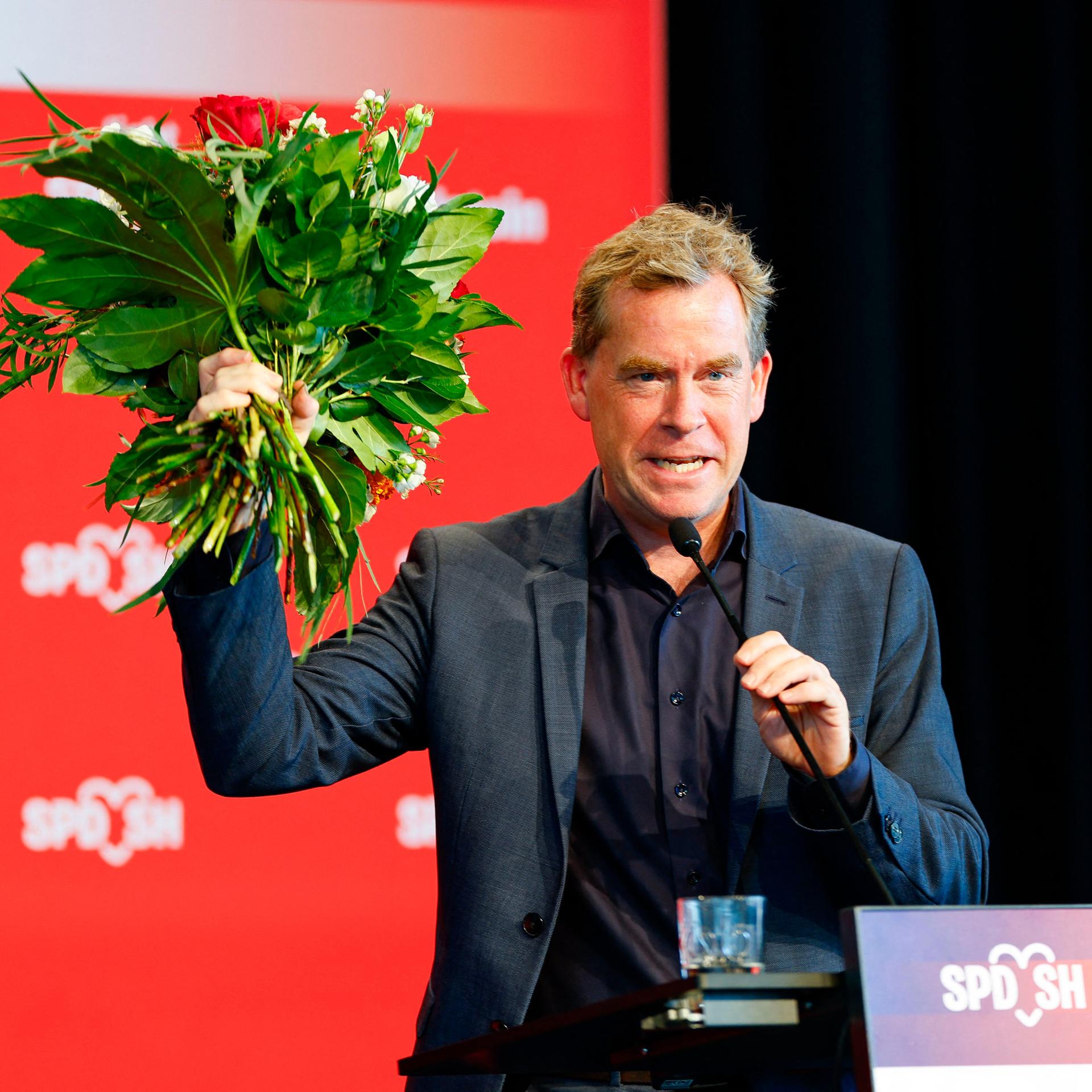 Ulf Kämpfer jubelt mit einem Blumenstrauß in der Hand auf einer SPD-Veranstaltung.